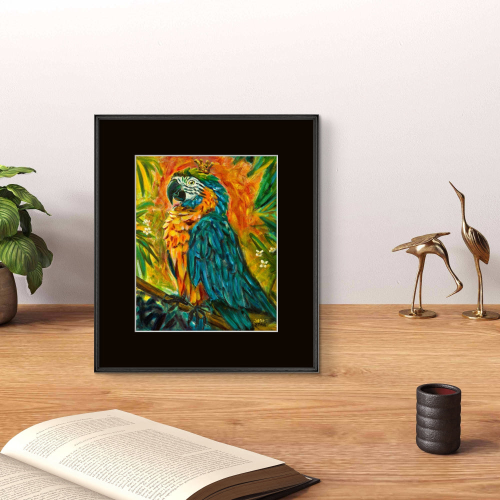 A framed colorful painting of a blue-and-yellow macaw perched on a branch, displayed on a wooden tabletop against a light wall, with a plant, book, and two gold bird figurines nearby.