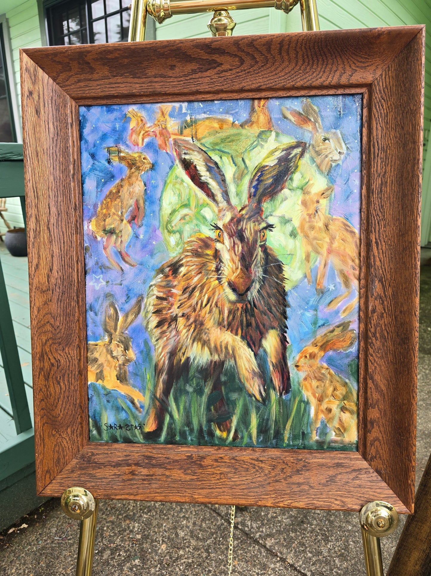 A framed painting of hares and rabbits on an easel. The artwork features a large hare in the center with smaller hares around it, painted in vivid blue, green, brown, and orange tones.