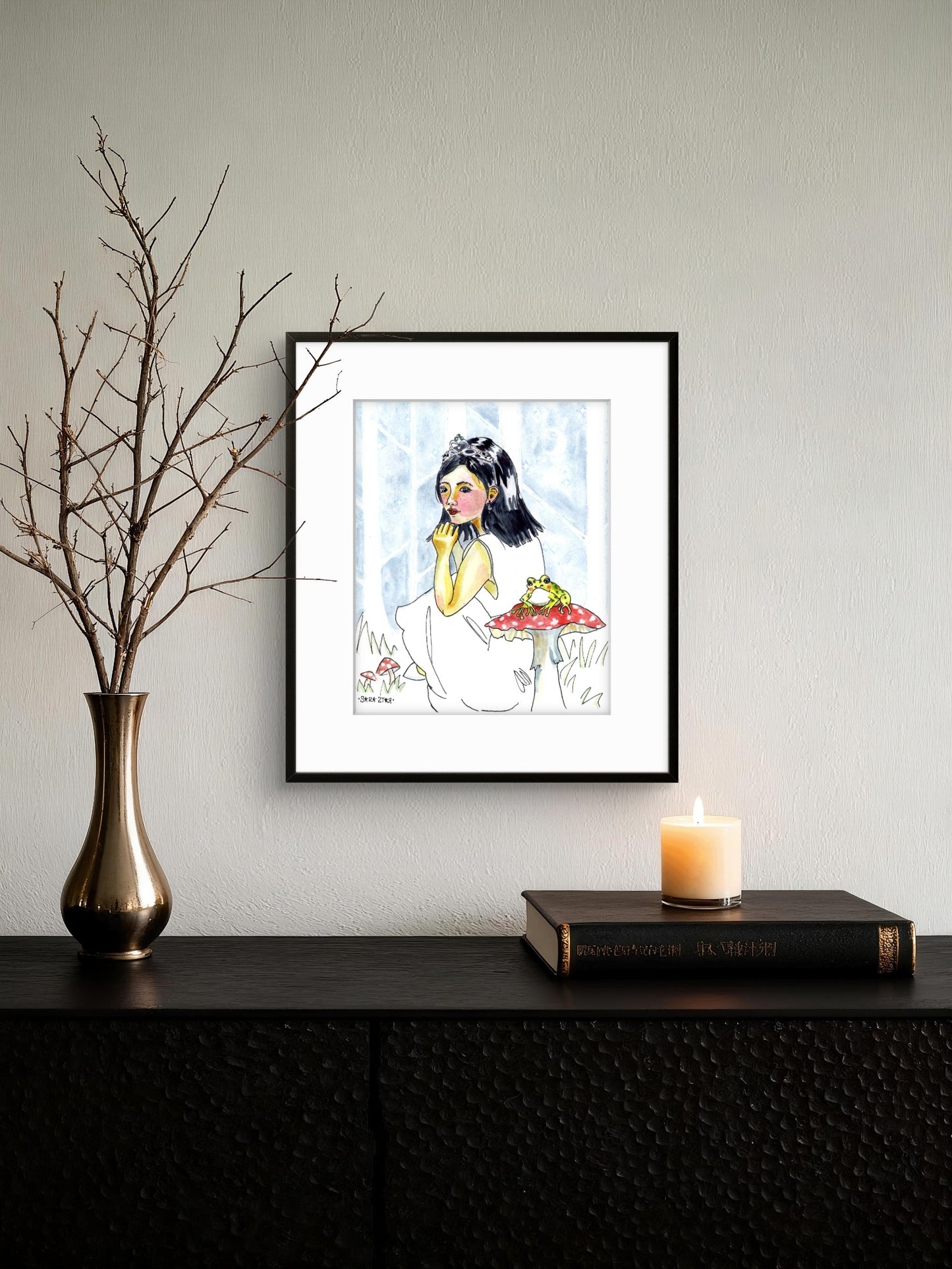 A framed art print featuring a watercolor-style illustration of a girl in a white dress holding a frog, displayed on a wall above a dark console table with a vase, candle, and book.
