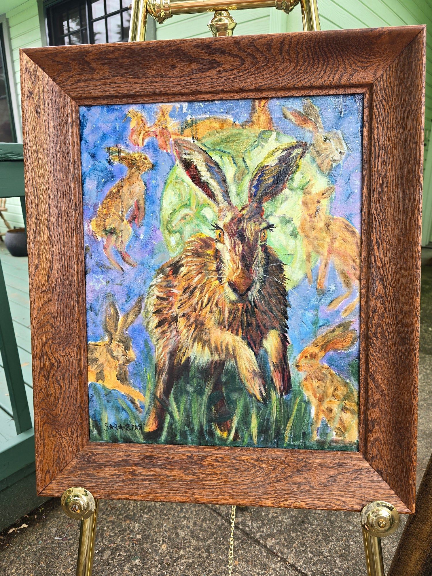 A framed painting of hares and rabbits on an easel. The artwork features a large hare in the center with smaller hares around it, painted in vivid blue, green, brown, and orange tones.
