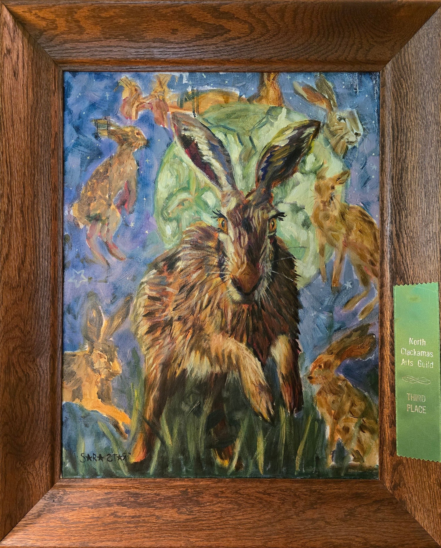 A framed painting depicting a large brown hare in the center with several smaller hares around it against a blue and green background. The artwork is housed in a thick dark wood frame, and a green ribbon is attached to the right side.