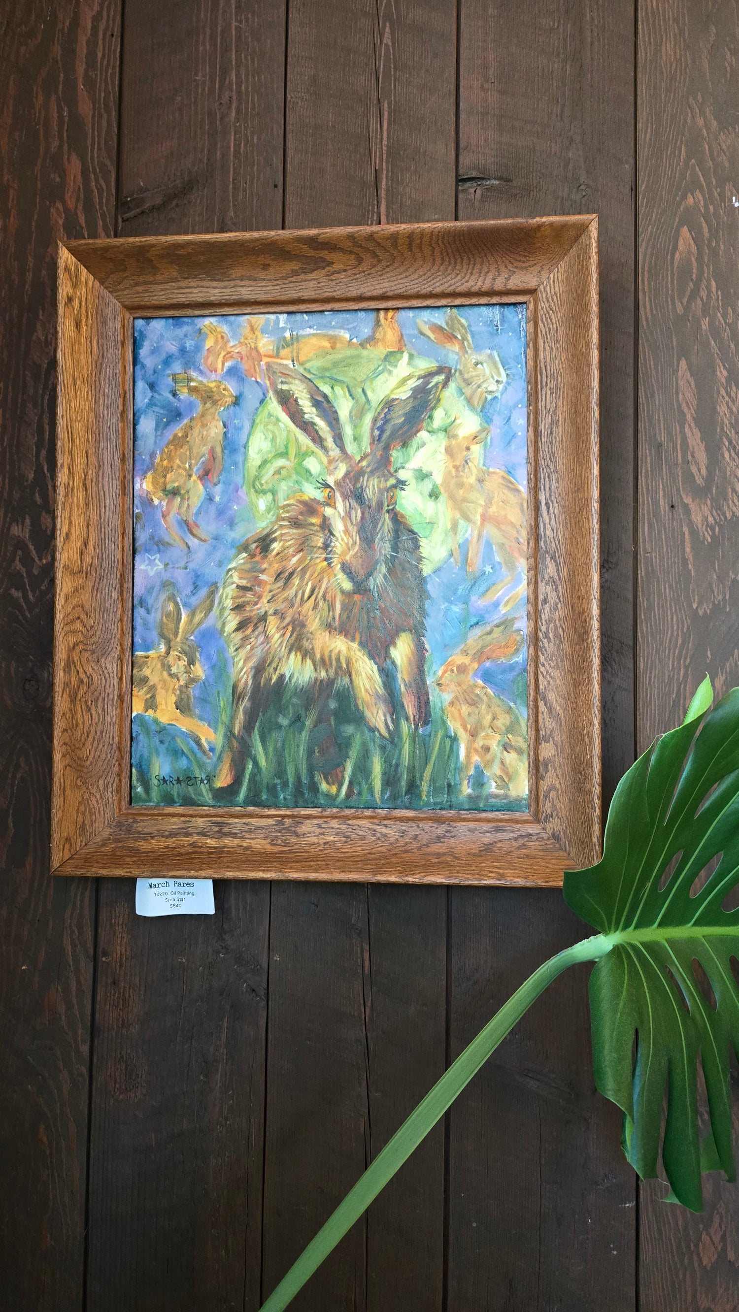 A framed painting of a hare and other hares in a grassy field, displayed on a dark wood plank wall with a green plant leaf in the foreground.