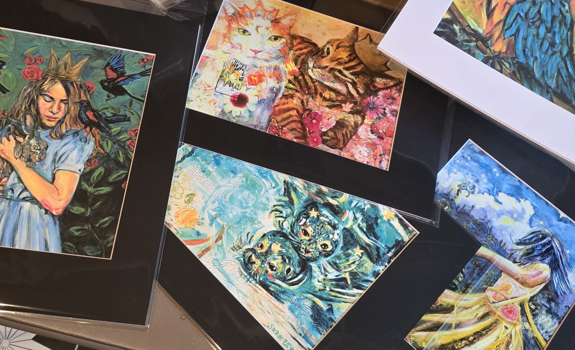 Several colorful art prints with black mats are arranged on a tabletop. The prints feature whimsical illustrations including a girl in a blue dress, a tiger with flowers, swirling blue abstract forms, and a woman in a yellow dress.