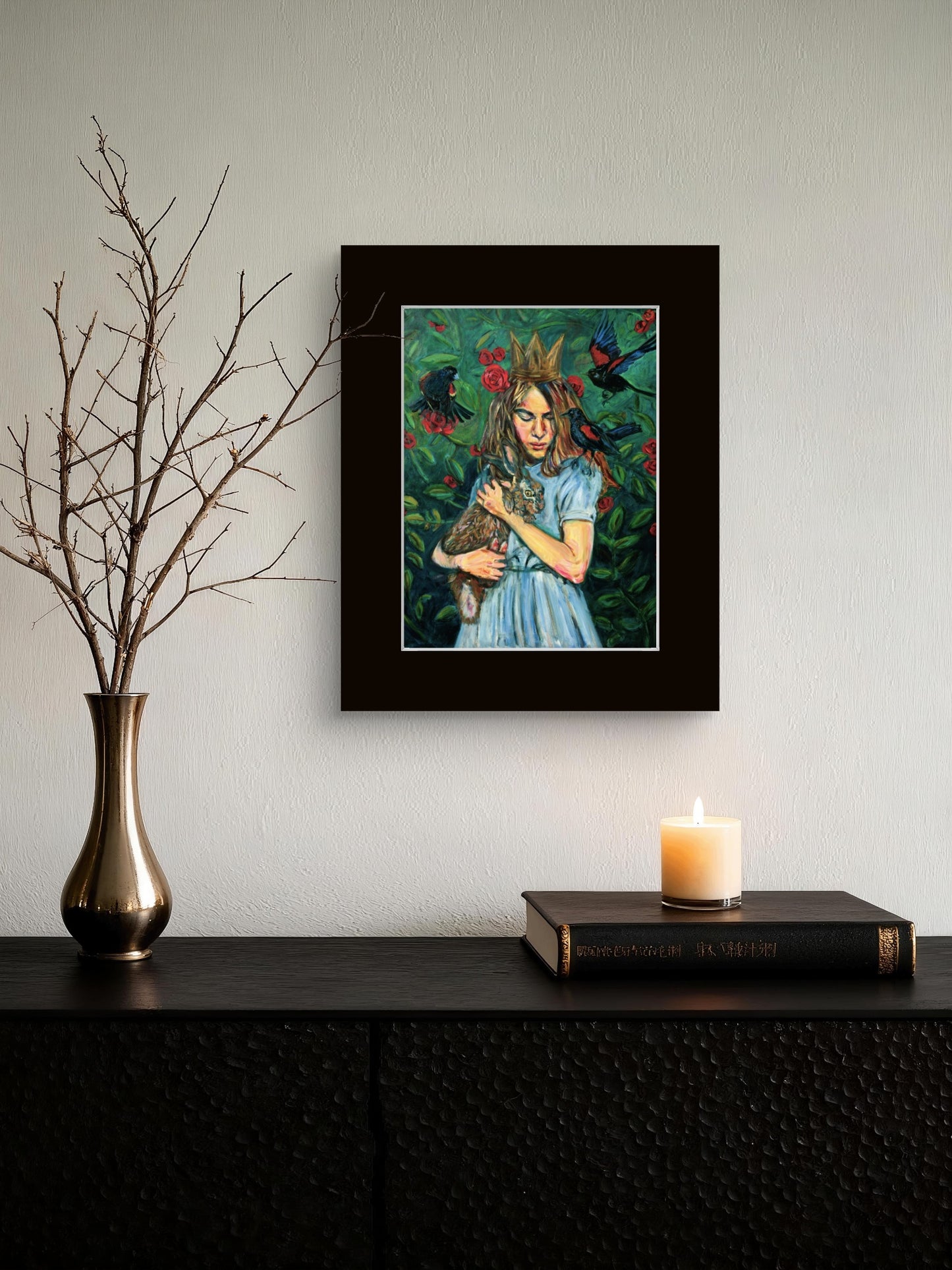 A framed art print depicting a woman holding a rabbit, hung on a light gray wall above a dark console table with a vase, candle, and book.