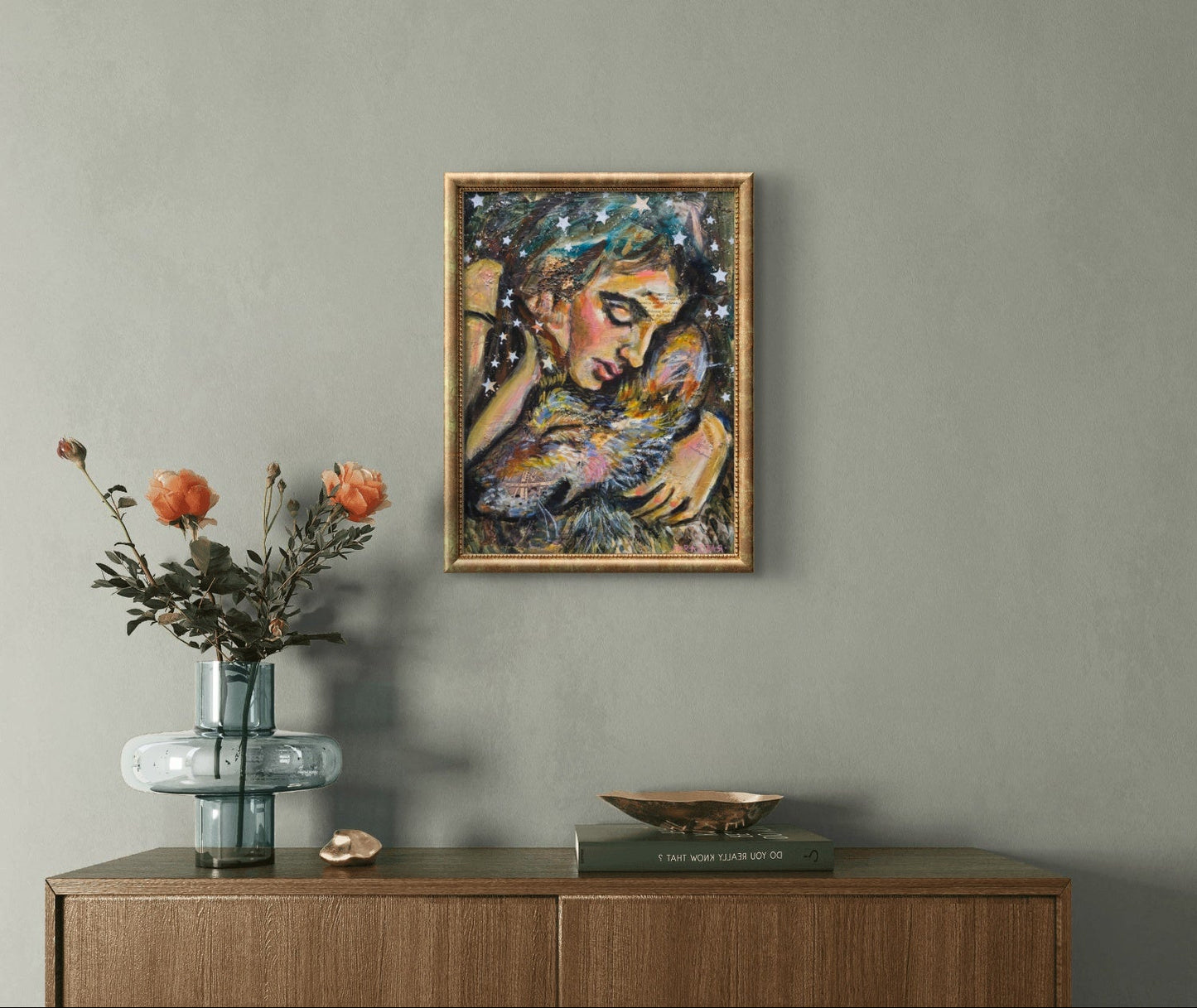 A framed abstract painting of a woman holding a wolf on a textured gray wall above a wooden console table with a glass vase of orange flowers and decorative objects.