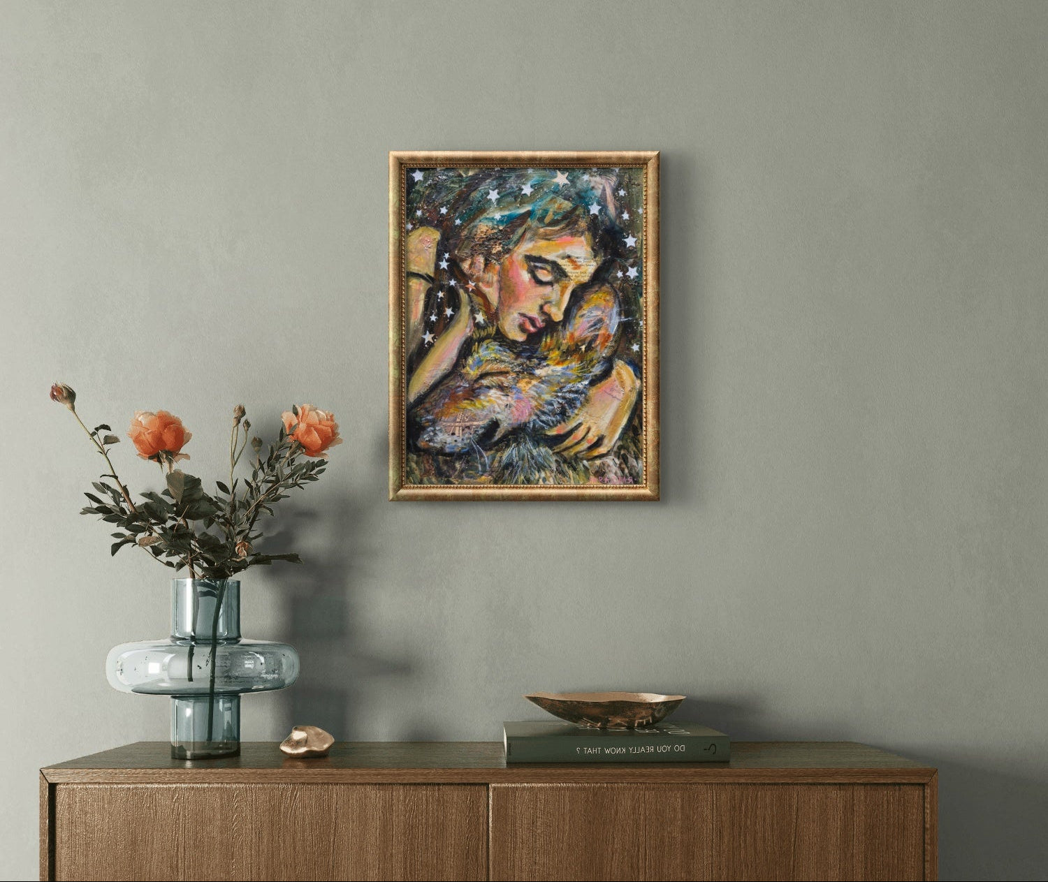 A framed abstract painting of a woman holding a wolf on a textured gray wall above a wooden console table with a glass vase of orange flowers and decorative objects.